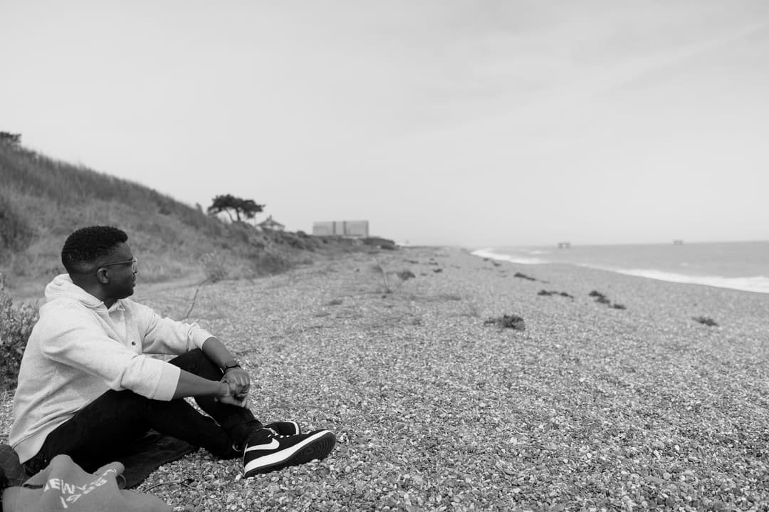 man sat down on a stoney beach with legs crossed looking away from the camera, deep in thought