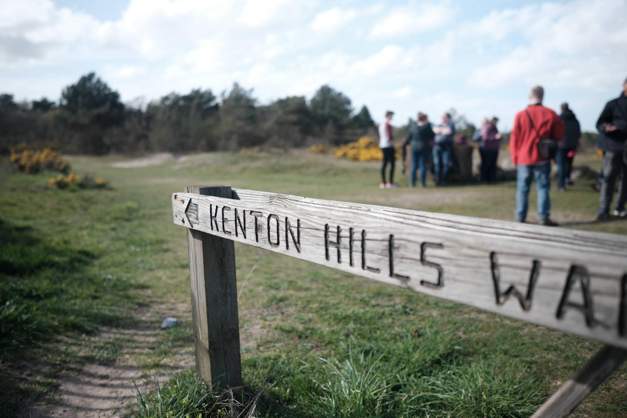 group of people standing behind an in-focus, slighty worn, wooden sign pointing towards 'Kenton Hills'