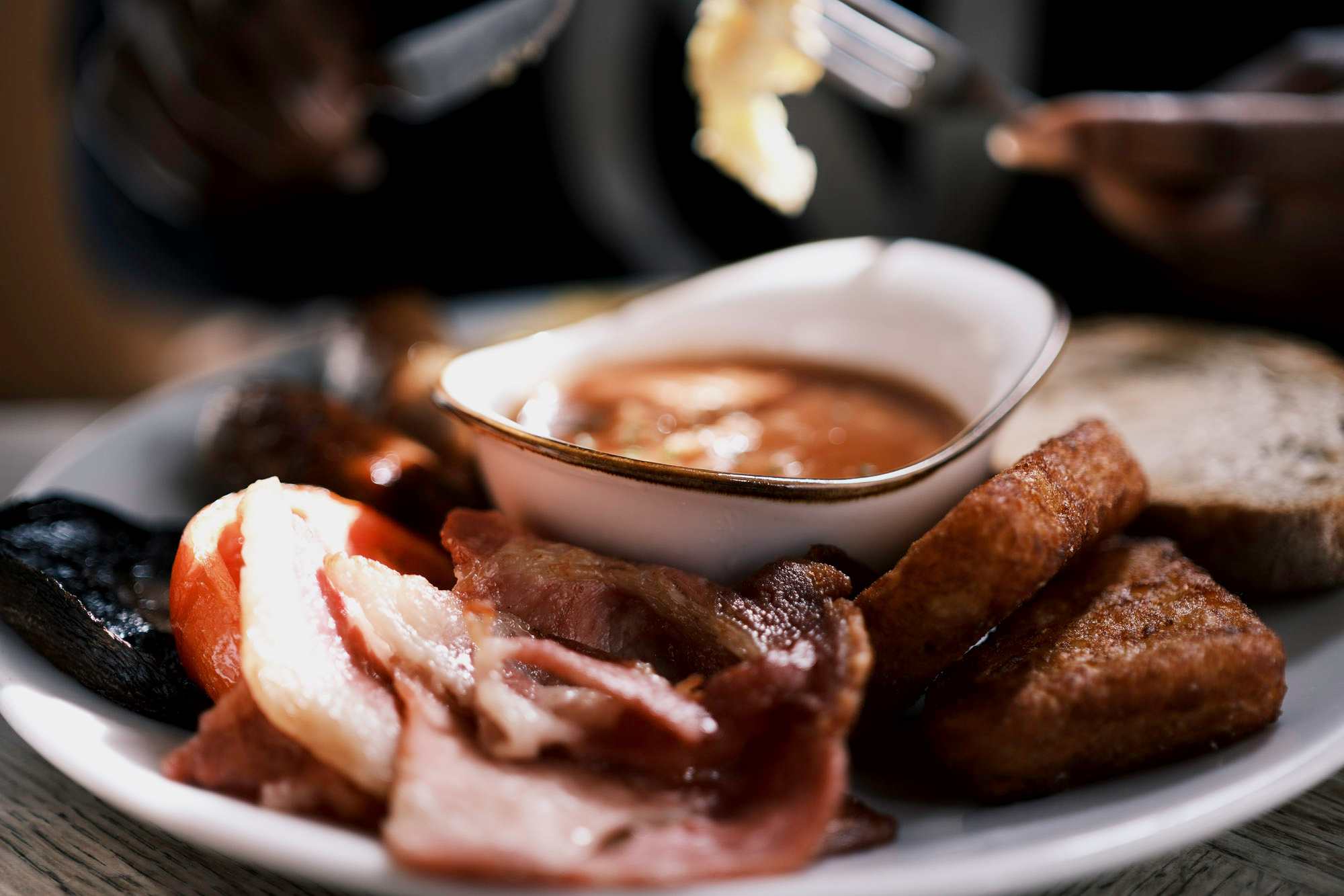 full english breakfast of fluffy eggs, fried bacon, crispy hash browns, mushrooms, tomatoes, thick sausages, toast and a bowl of beans, presented nicely on a plate.
