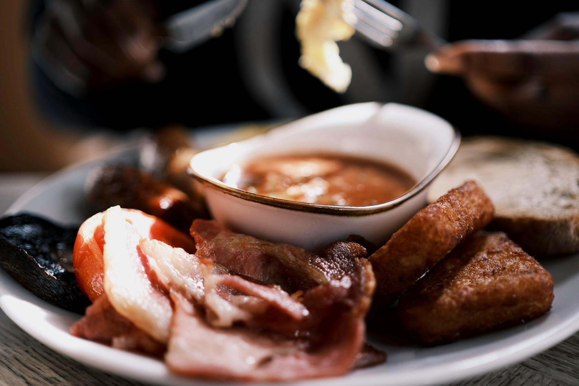full english breakfast of fluffy eggs, fried bacon, crispy hash browns, mushrooms, tomatoes, thick sausages, toast and a bowl of beans, presented nicely on a plate.