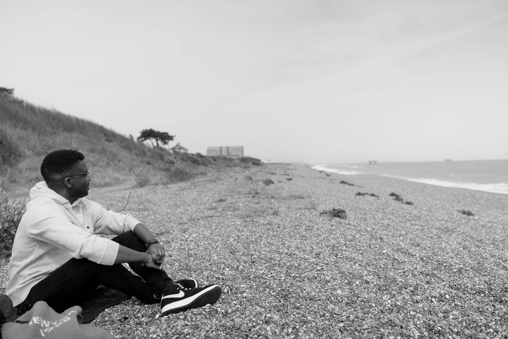 man sat down on a stoney beach with legs crossed looking away from the camera, deep in thought